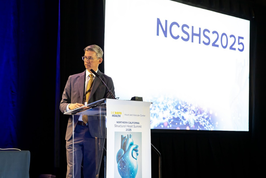 A speaker presents at a podium during a conference session with a presentation screen displayed in the background.